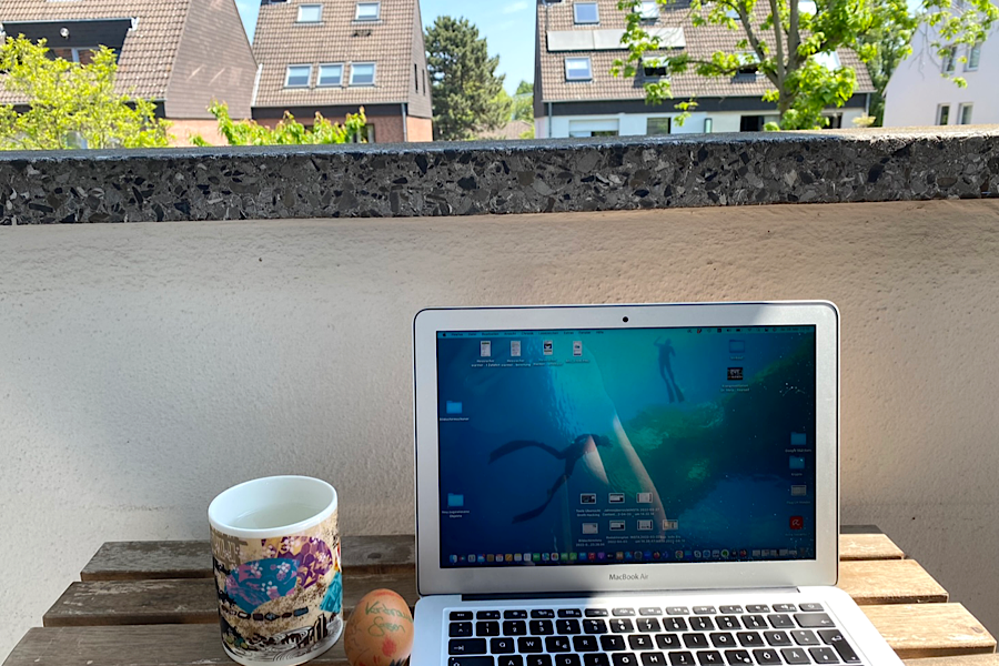 Bild eines aufgeklappten Laptop auf einem Holztisch auf dem Balkon mit Blick ins Grüne. Daneben liegt ein dekoriertes Ei und es steht eine Tasse mit Blumenmotiv.