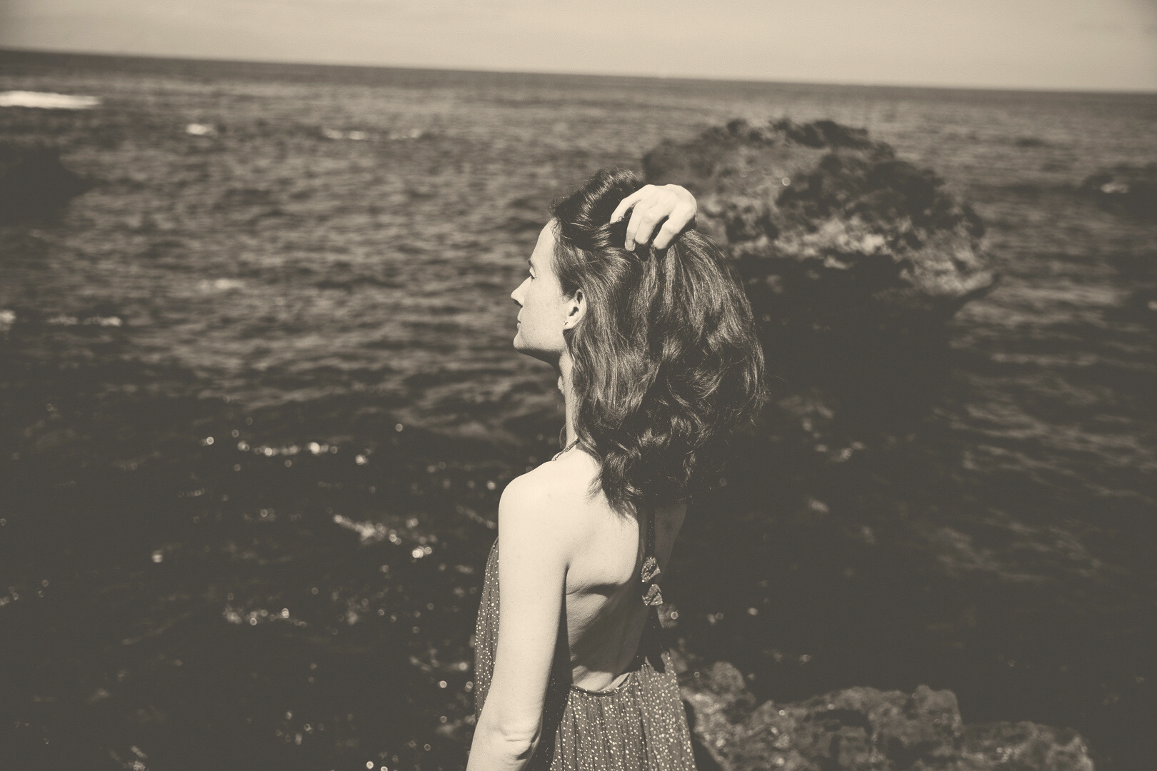 Schwarz weiß Bild mit leichtem Braunton aufbereitet. Eine Frau blickt aufs Meer, man sieht sie von der Seite. Sie hat eine Hand am hinteren Kopf und wirkt nachdenklich