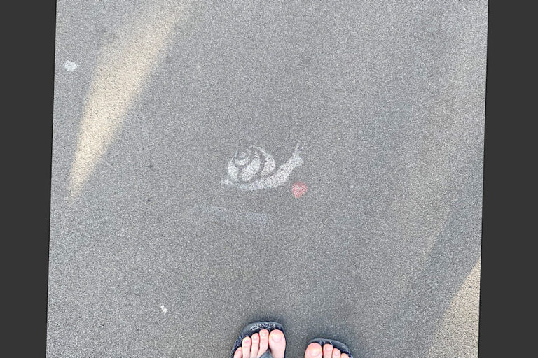 Abbildung einer aufgemalten Schnecke mit Herz davor auf Beton. Im unteren Bildrand sieht man Füße in Sandalen