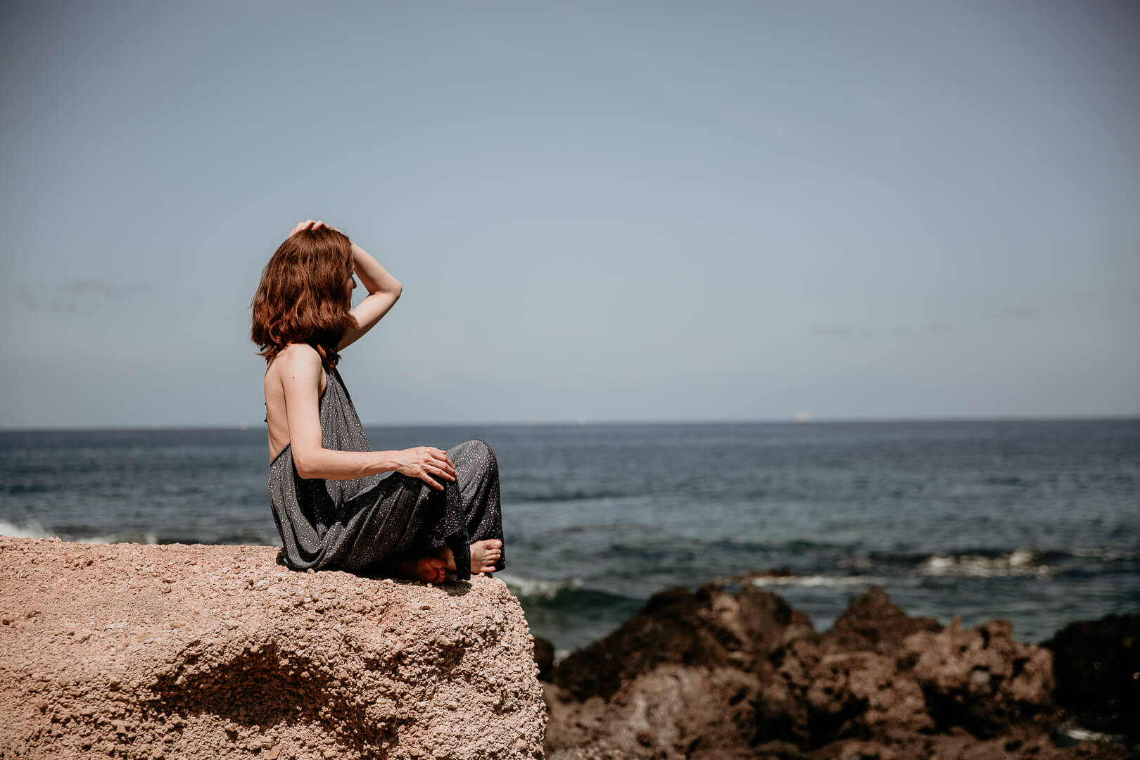 Frau sitzt auf Felsen am Meer, hat eine Hand an der Seite am Kopf im Haar.