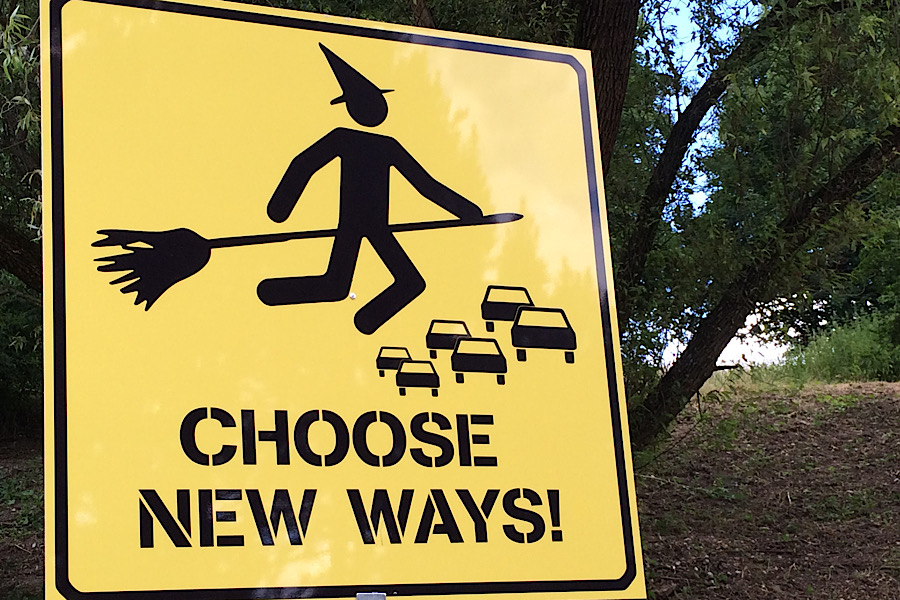 Ein gelbes Schild, wie ein Verkehrsschild mit dem Schriftzug "Choose new ways!". Abgebildet ist eine stilisierte Hexe auf einem Besen, die über einen Verkehrsstau mit Autos fliegt.